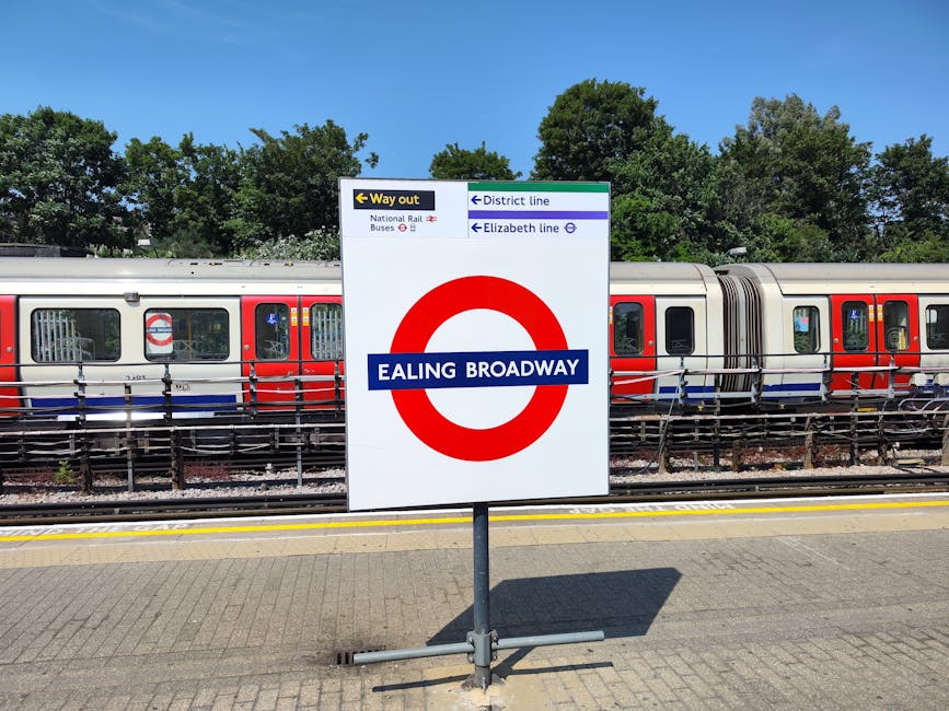 A large white sign with a red London Underground roundel logo displaying the name Ealing Broadway in blue, positioned outdoors on a paved platform near train tracks. Behind the sign, a modern train wi