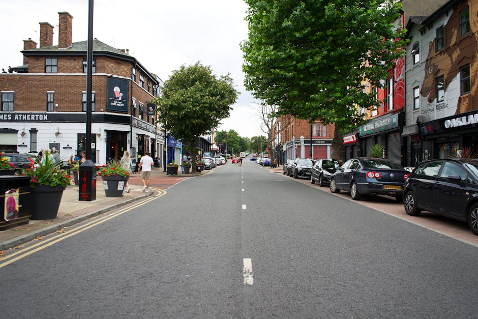 A wide urban street scene during daytime showing the loading process of a house or commercial move by [COMPANY_NAME], with parked cars lining both sides of the road. On the left, there are potted plan