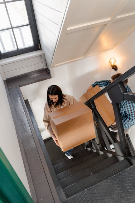 Inside a residential property, a woman with long dark hair, dressed in a beige sweater, is carrying a large, sealed cardboard box up a staircase. The staircase has dark carpeted steps with wooden edge